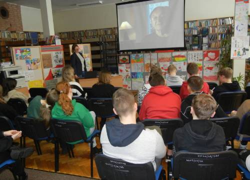 Widok od tyłu na młodzież uczestniczącą w zajęciach w bibliotece, w tle widać prowadzącą dr Barbarę Męczykowską oraz ekran z prezentacją.